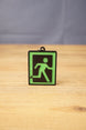 Tarkov inspired extraction keyring standing upright on a wooden tabletop, featuring a green running figure sign with black background and frame.