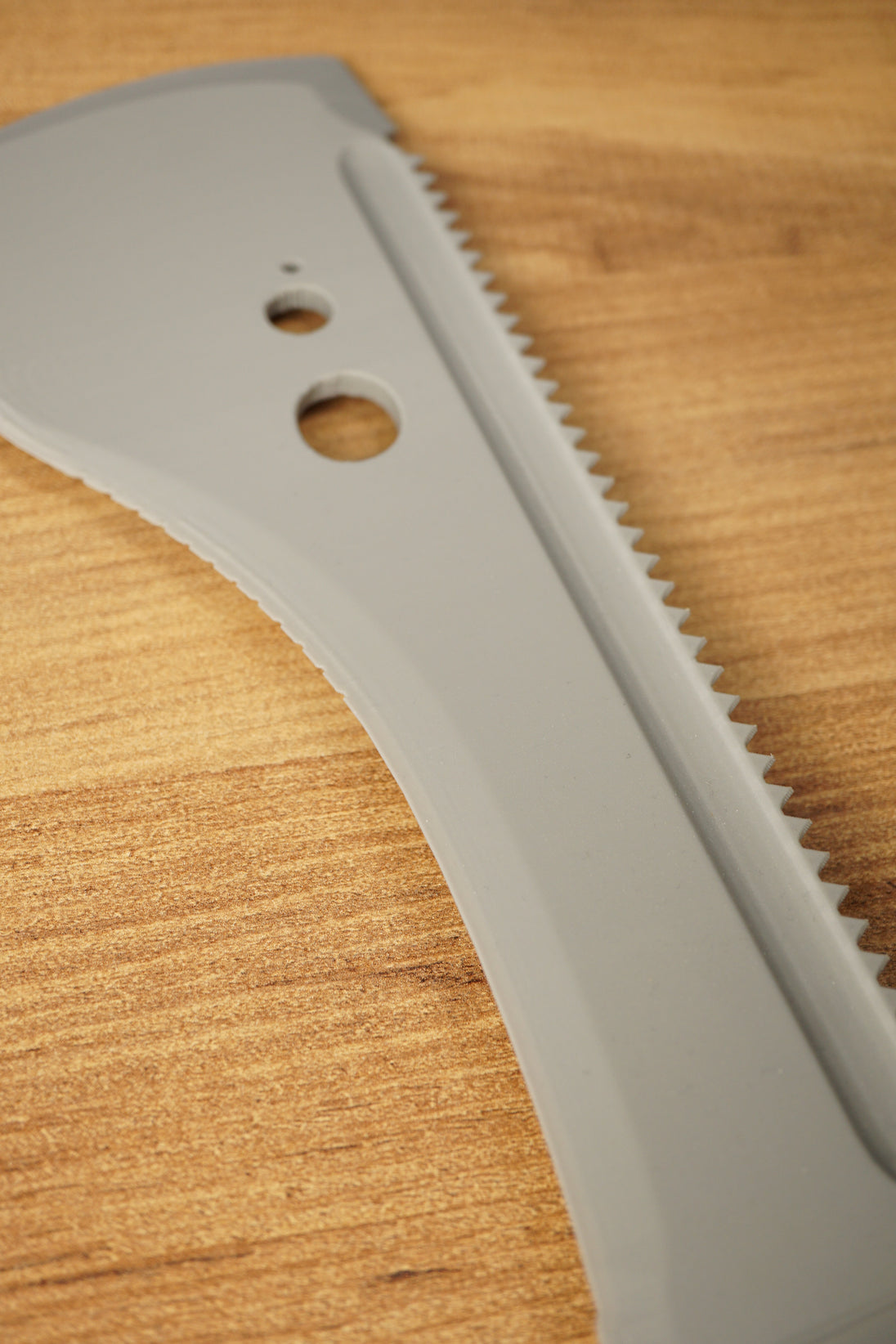 Macro close-up of the gray 3D-printed machete blade on a wooden surface, showing serrated teeth and circular cutouts.