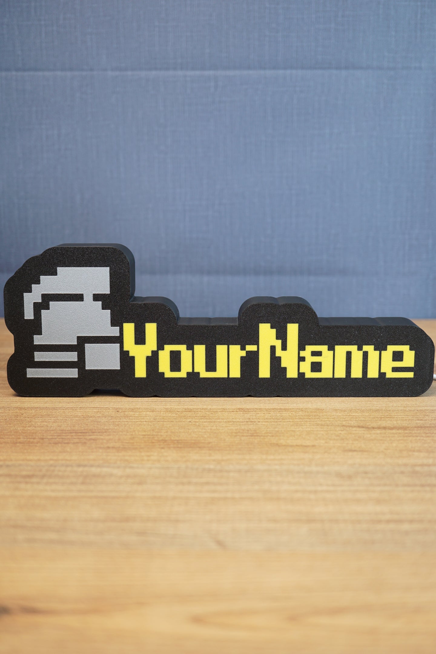 Ultimate ironman RuneScape-style nameplate with a silver helmet icon, bright yellow custom text, and black border displayed on a desk.