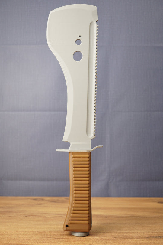 Full upright view of a 3D-printed UVSR Taiga-1 survival machete replica standing on a wooden table, featuring a gray blade and brown ribbed handle.