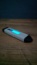 Glow in the dark WARDOGS Adrenaline Pen replica viewed horizontally in dim lighting, displaying its luminous blue panel and white ribbed casing.