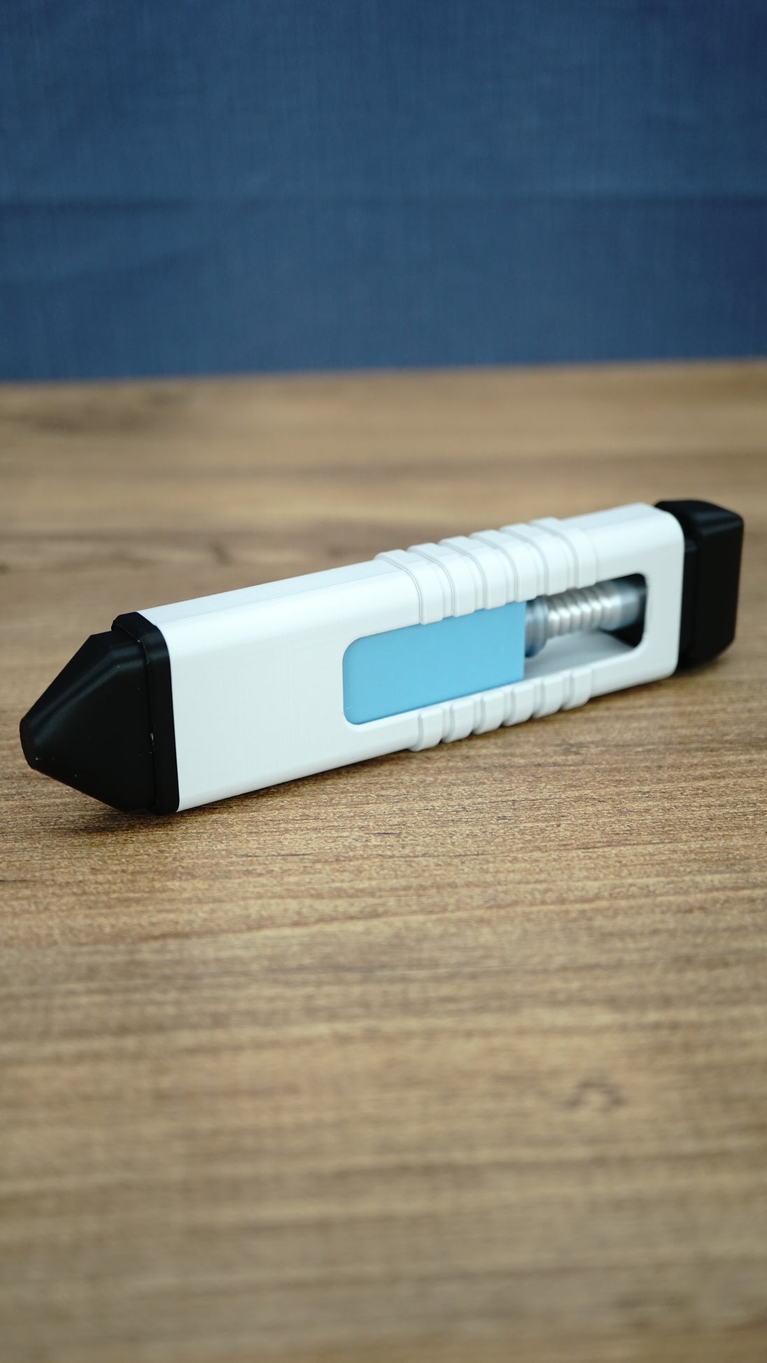 Side view of the 16cm WARDOGS Adrenaline Pen clicker fidget toy resting on a wooden tabletop, showing its blue insert, silver mechanism and black ends.