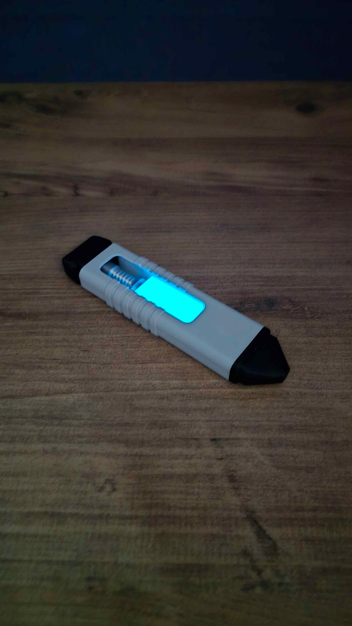 WARDOGS Adrenaline Pen glow in the dark fidget toy lying diagonally on a wooden table with its blue central panel glowing bright cyan in low light.