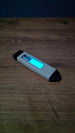 WARDOGS Adrenaline Pen glow in the dark fidget toy lying diagonally on a wooden table with its blue central panel glowing bright cyan in low light.