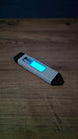 WARDOGS Adrenaline Pen glow in the dark fidget toy lying diagonally on a wooden table with its blue central panel glowing bright cyan in low light.