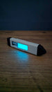 WARDOGS Adrenaline Pen fidget clicker with bright blue glow in the dark centre, casting a cyan reflection onto the wooden tabletop beside it.