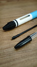 Close-up of a black ballpoint refill and transparent pen next to the white and blue WARDOGS Adrenaline Pen replica on a wooden surface.