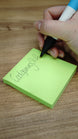 Hand using the WARDOGS Adrenaline Stim Pen replica to write on a green sticky note, demonstrating its functional ballpoint writing tip.