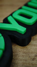 Macro close-up of the Manticore Green gaming nameplate showing bright green raised lettering, its black backing and the detailed layered construction.