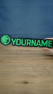 Manticore Green 25cm gaming nameplate held in one hand, showing the green lion emblem, YOURNAME lettering and full black outlined design against a blue background.