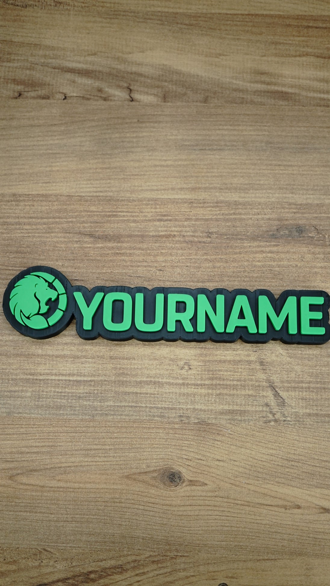 Overhead product photograph of the Manticore Green nameplate with bright green YOURNAME lettering and a circular lion faction emblem on black backing.