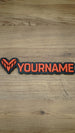 Overhead photograph of a Valkyra Red gaming nameplate featuring an angular red-orange faction emblem and YOURNAME lettering on a black backing.