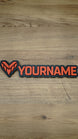 Overhead photograph of a Valkyra Red gaming nameplate featuring an angular red-orange faction emblem and YOURNAME lettering on a black backing.