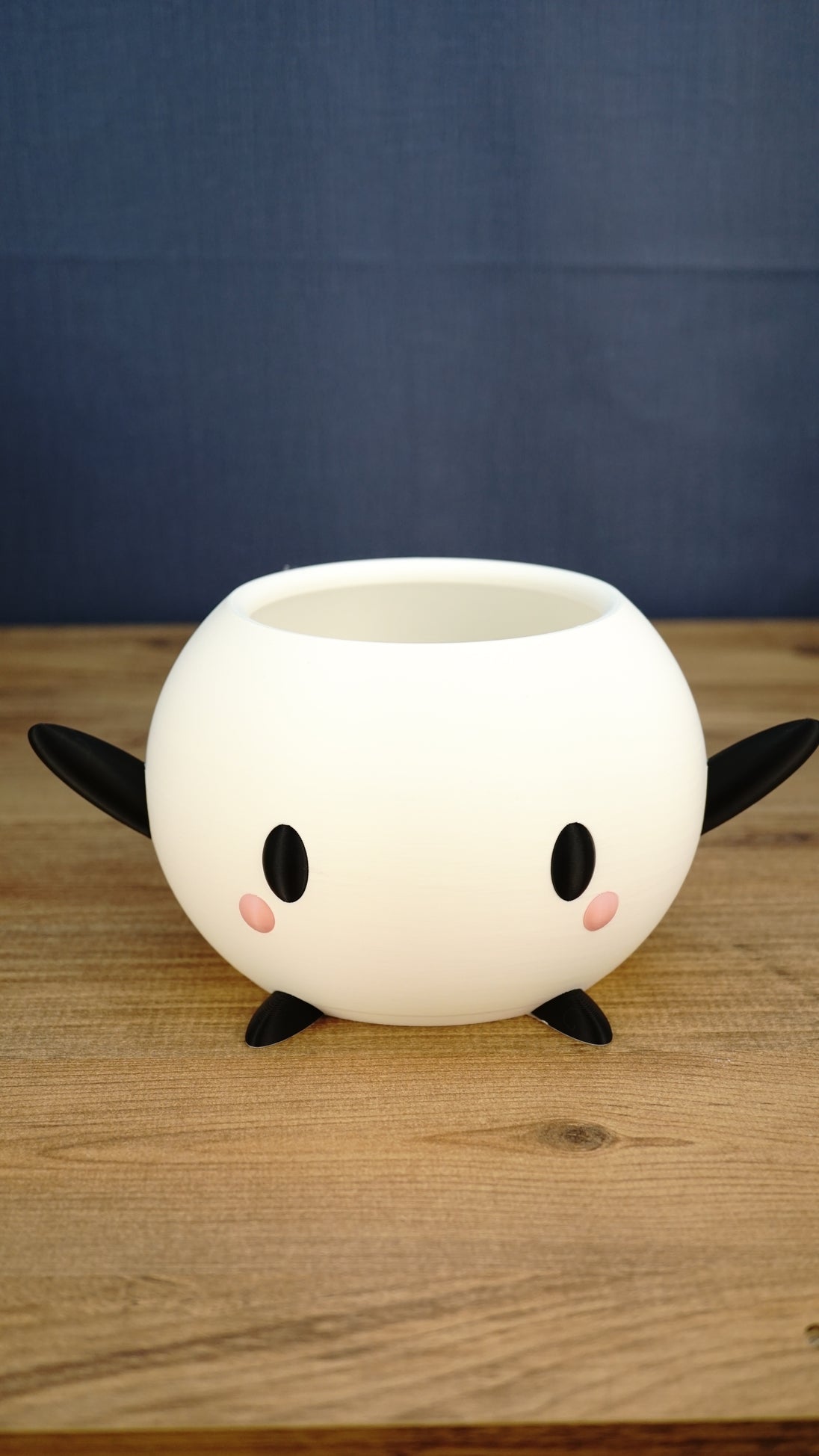 White Stardew Valley-inspired Junimo planter pot with black arms and feet, oval eyes, and pink cheeks on a wooden tabletop.
