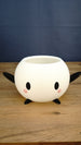 White Stardew Valley-inspired Junimo planter pot with black arms and feet, oval eyes, and pink cheeks on a wooden tabletop.