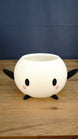White Stardew Valley-inspired Junimo planter pot with black arms and feet, oval eyes, and pink cheeks on a wooden tabletop.
