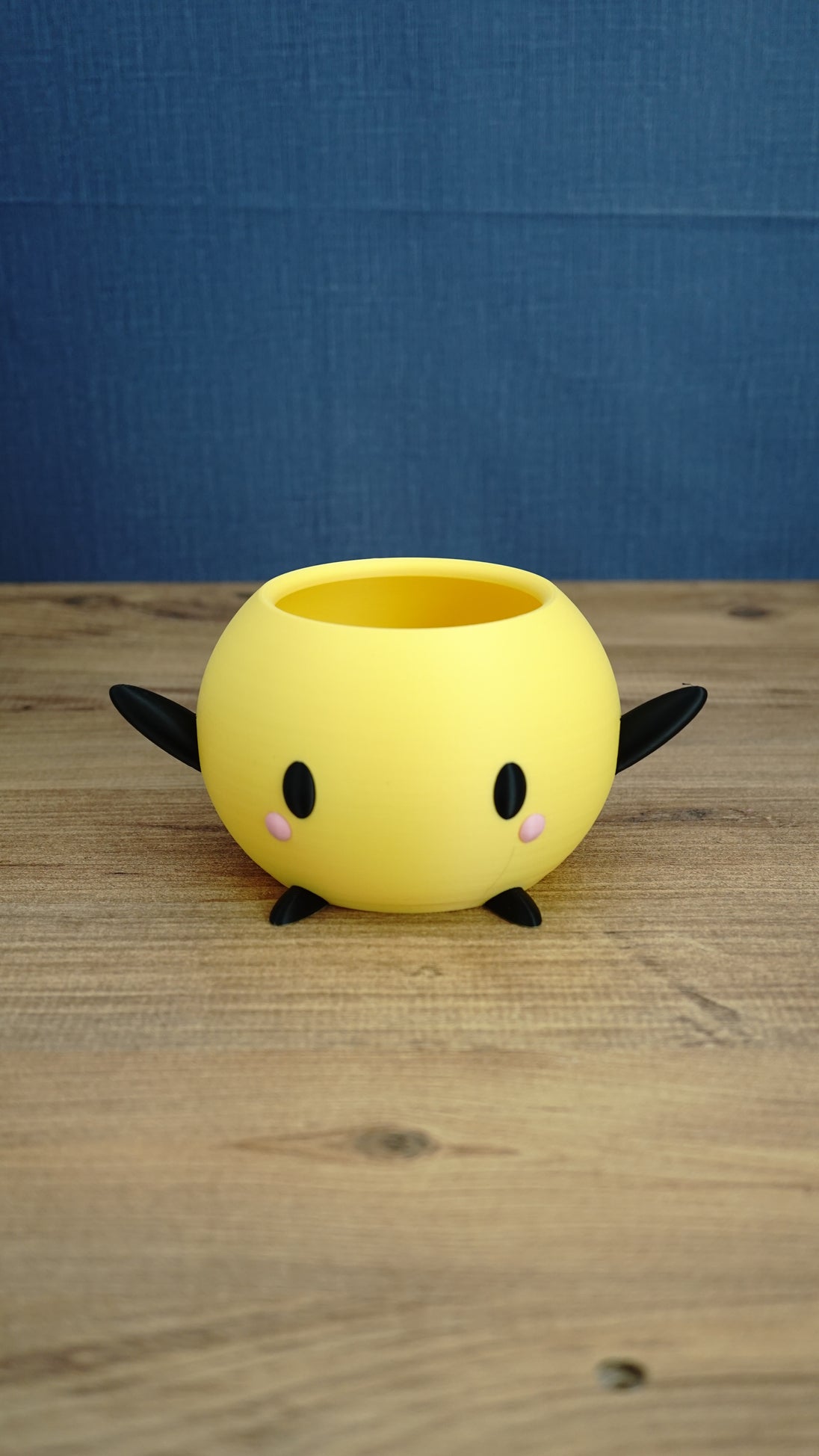 Yellow Stardew Valley Junimo planter pot with black arms, black feet, oval eyes, and pink cheeks on a wooden tabletop.