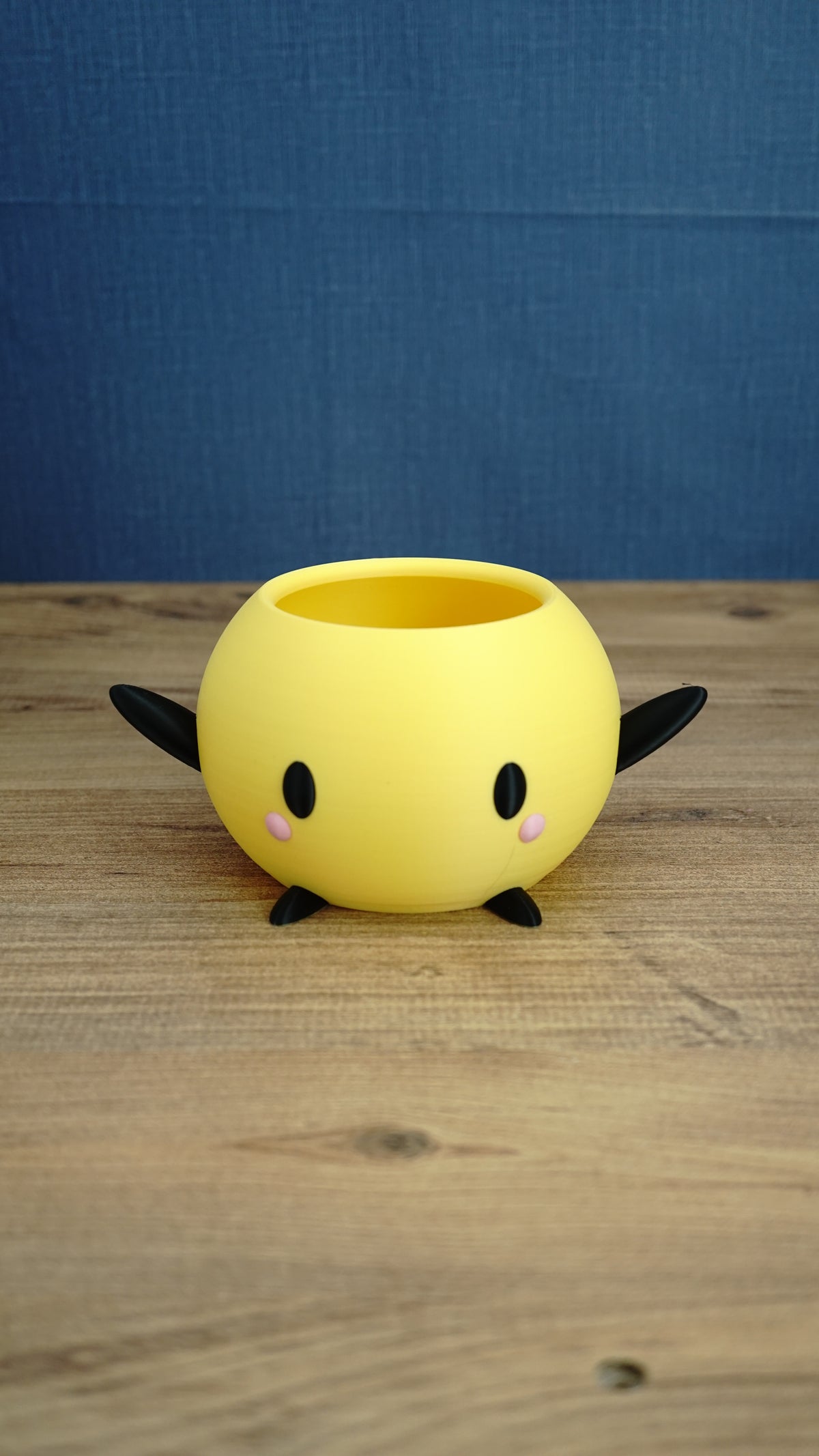 Yellow Stardew Valley Junimo planter pot with black arms, black feet, oval eyes, and pink cheeks on a wooden tabletop.
