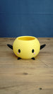 Yellow Stardew Valley Junimo planter pot with black arms, black feet, oval eyes, and pink cheeks on a wooden tabletop.
