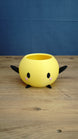 Yellow Stardew Valley Junimo planter pot with black arms, black feet, oval eyes, and pink cheeks on a wooden tabletop.